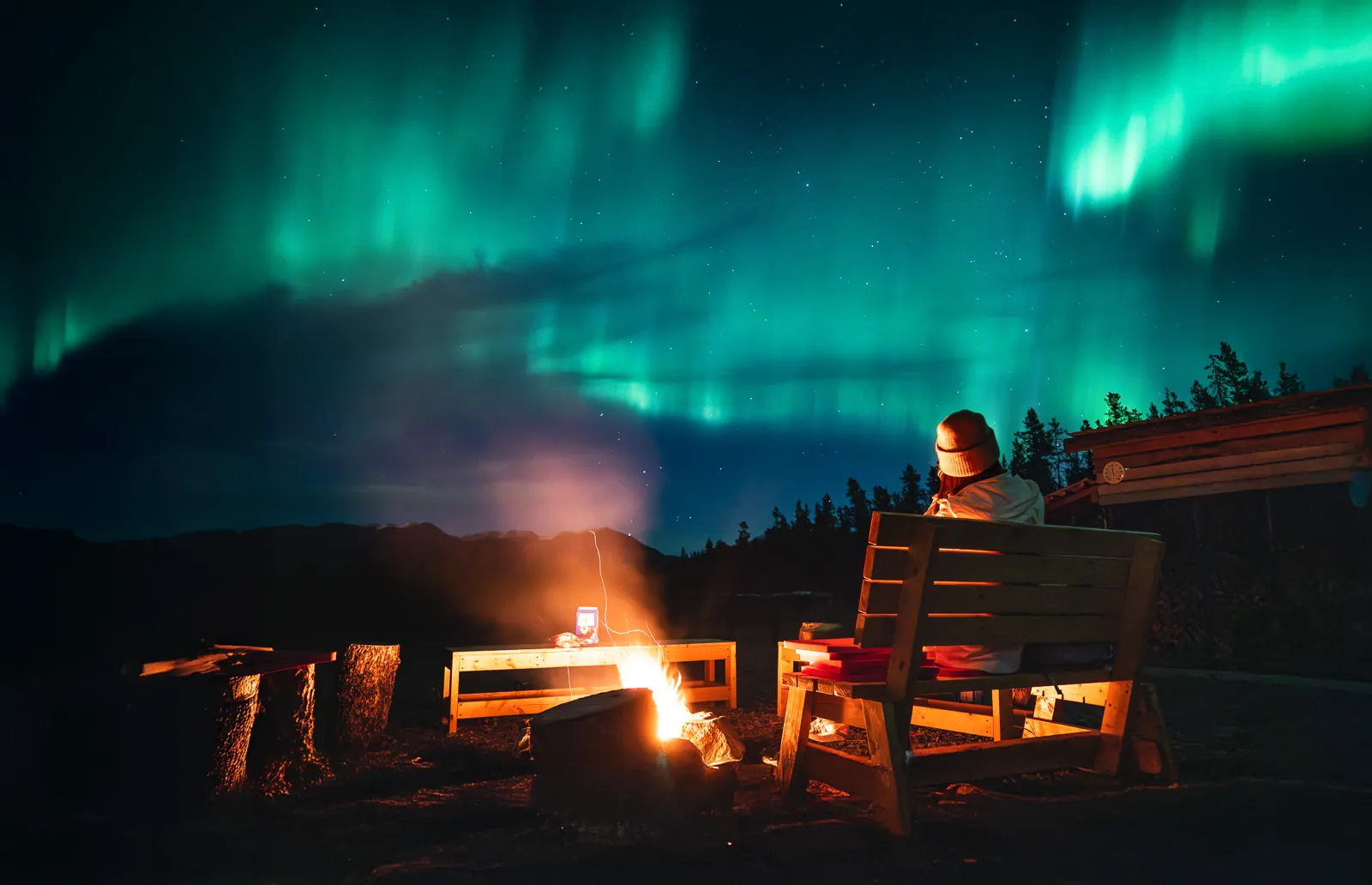 A guest beside the outdoor campfire at the Aurora Lounge as green northern lights sweep overhead.