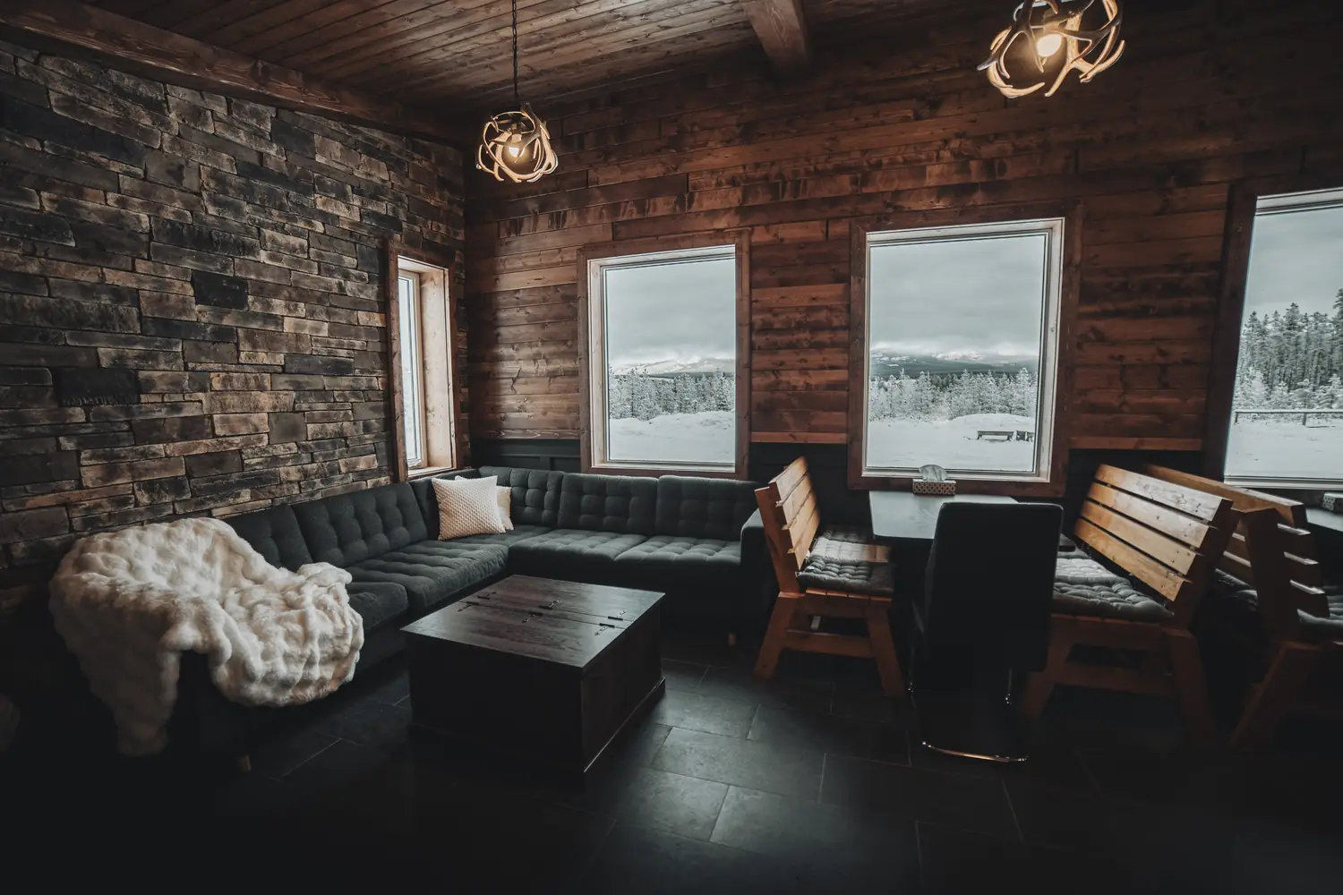 Charcoal sectional sofa with a white fur throw beneath warm pendant lights, with snowy forest visible through the windows