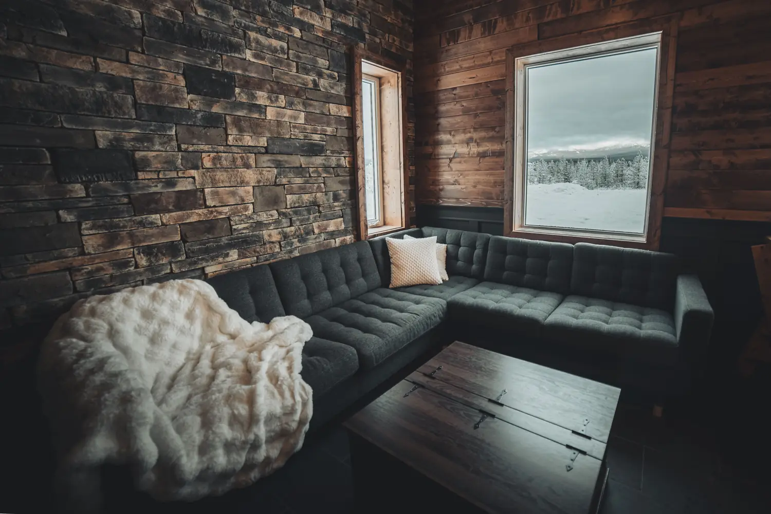 Corner sectional with a white fur blanket beside the stacked-stone wall and snowy window.