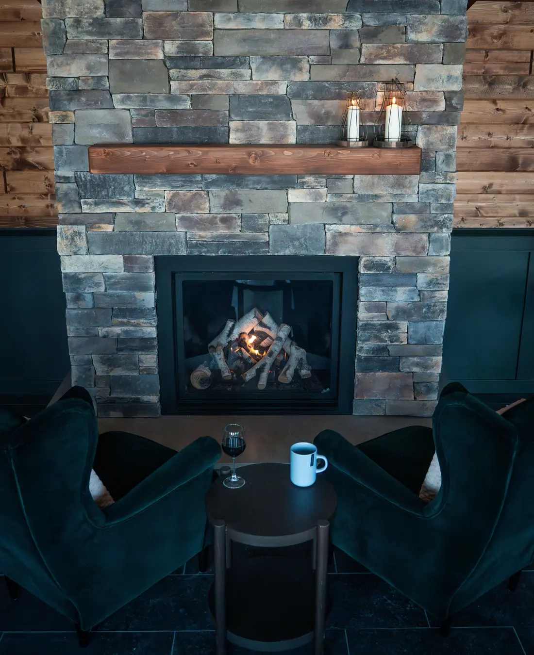 Two deep-green armchairs facing the stone fireplace, with red wine and a hot drink on the table between them.